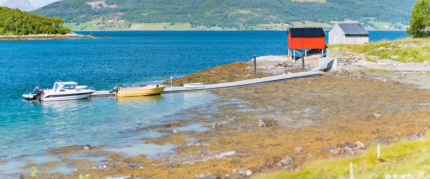 Beautiful landscape of Norway. Red barn, trees and water, mountains in background. Travel in Scandinavia, Europe. Boats at pier.