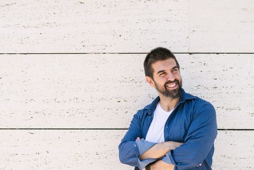 Bearded male leaning on wall with crossed arms while looking away with a smile