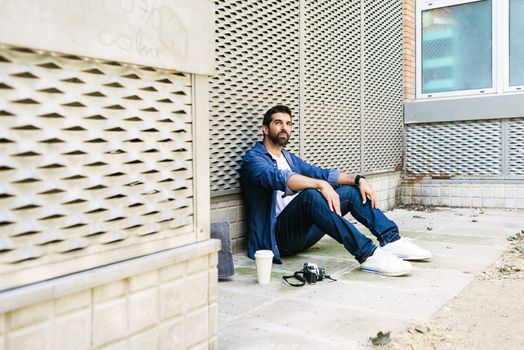 Cheerful bearded man in casual attire sitting on street ground while relaxing on a coffee break