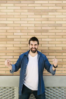 Joyful young bearded male in casual blue shirt posing on bricked wall background.