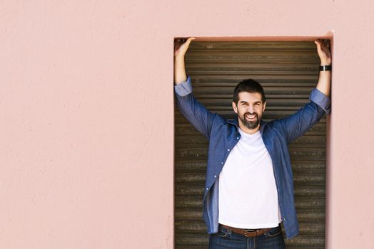 Bearded male standing against pink wall with arms raised while looking camera