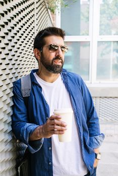 Portrait of a handsome man in blue t-shirt standing with coffee to go on a metallic wall background