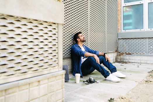 Cheerful bearded man in casual attire sitting on street ground while relaxing on a coffee break