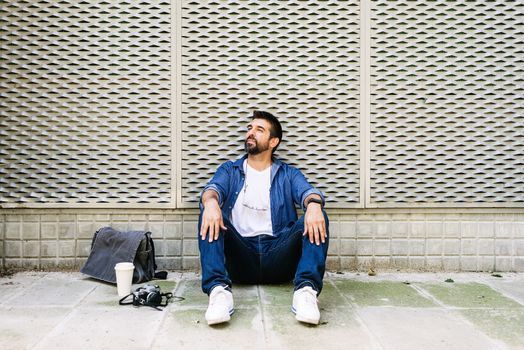 Cheerful bearded man in casual attire sitting on street ground while relaxing on a coffee break