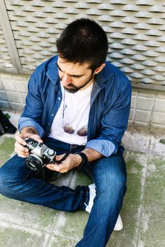 Cheerful bearded traveler man sitting on ground while using checking a camera