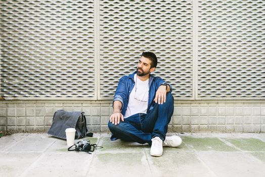 Cheerful bearded man in casual attire sitting on street ground while relaxing on a coffee break