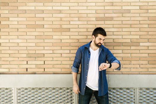 Bearded male looking at his watch against a bricked wall outdoors