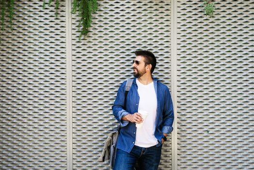 Portrait of a handsome man in blue t-shirt standing with coffee to go on a metallic wall background