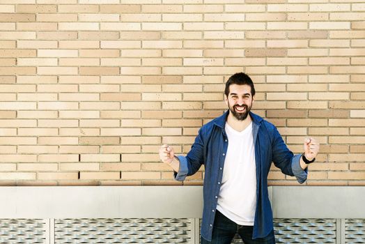 Joyful young bearded male in casual blue shirt posing on bricked wall background.
