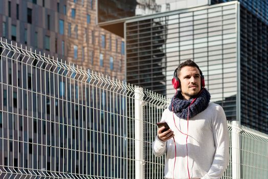Young bearded male with headphones and holding smartphone while walking against skyscrapers in sunny day