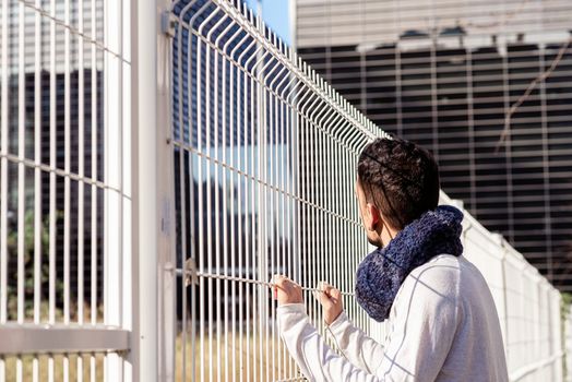 Young male leaning on a metallic fence while looking away to the business buildings