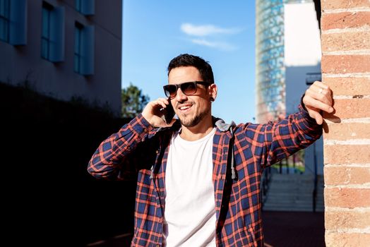 Young bearded male leaning on a bricked wall wearing sunglasses while using a smartphone outside.