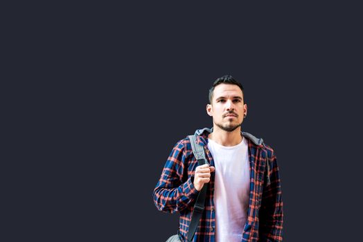 Young bearded guy leaning on a dark wall holding a shoulder bag while looking away