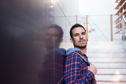 Young bearded guy leaning on a dark wall holding a shoulder bag while looking away