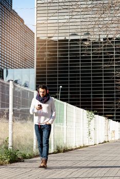 Young bearded male with headphones and holding smartphone while walking against skyscrapers in sunny day