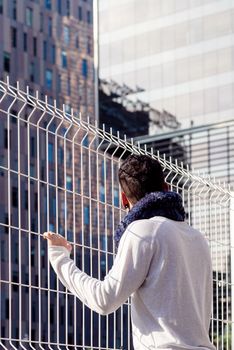 Young male leaning on a metallic fence while looking away to the business buildings