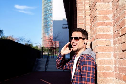 Young bearded male leaning on a bricked wall wearing sunglasses while using a smartphone outside.