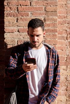 Young bearded man leaning on a bricked wall while using a smartphone