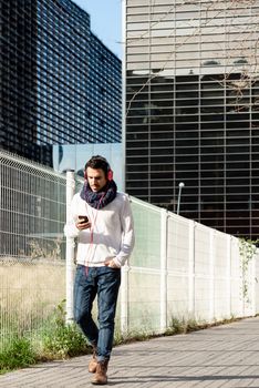 Young bearded male with headphones and holding smartphone while walking against skyscrapers in sunny day