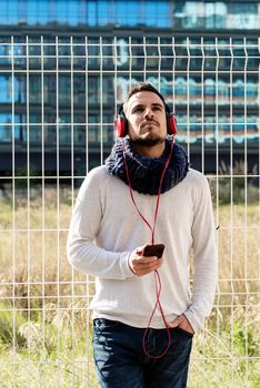 Young bearded man with headphones and holding smartphone while leaning on a metallic fence against skyscrapers in sunny day