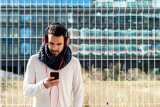 Young bearded man with headphones and holding smartphone while leaning on a metallic fence against skyscrapers in sunny day