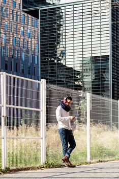 Young bearded man with headphones and holding smartphone while leaning on a metallic fence against skyscrapers in sunny day
