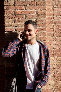 Young bearded male leaning on a bricked wall while using a smartphone outdoors.