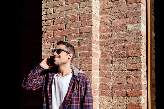 Young bearded male leaning on a bricked wall wearing sunglasses while using a smartphone outside.