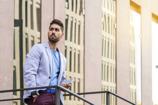 Portrait of a happy bearded young man holding a smartphone while standing outdoors and looking away