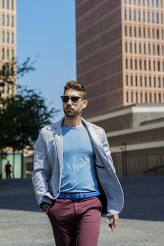 Portrait of a young bearded man with sunglasses, model of fashion, in urban background wearing casual clothes while walking with hand on pocket