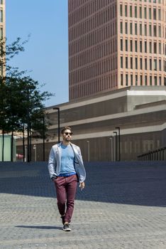 Portrait of a young bearded man with sunglasses, model of fashion, in urban background wearing casual clothes while walking with hand on pocket