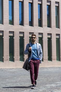 Young businessman walking next to office buildings while holding a shoulder bag outdoors