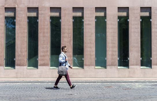 Young businessman walking next to office buildings while holding a shoulder bag outdoors