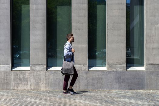 Young businessman walking next to office buildings while holding a shoulder bag outdoors