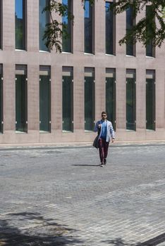 Young businessman walking next to office buildings while holding a shoulder bag outdoors