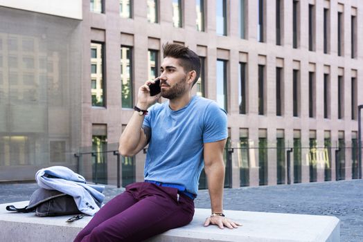 Portrait of cheerful modern businessman speaking by phone and smiling while sitting outdoors in a bench