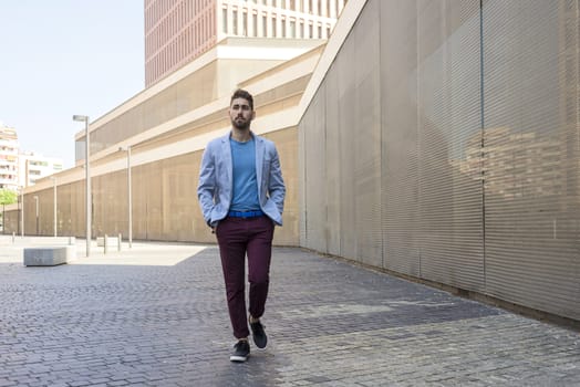 Portrait of a young bearded man, model of fashion, in urban background wearing casual clothes while walking with hands on pocket