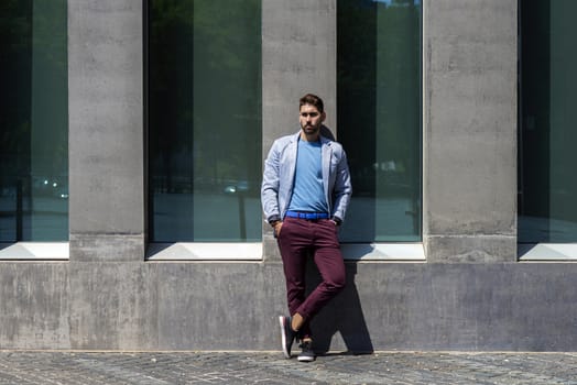 Portrait of a young bearded man, model of fashion, in urban background wearing casual clothes while leaning on a office building wall