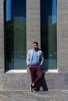 Portrait of a young bearded man, model of fashion, in urban background wearing casual clothes while leaning on a office building wall
