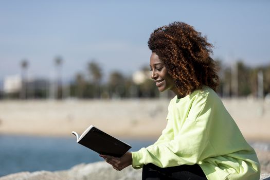 afro american woman sitting on shore while reading a book in a sunny day