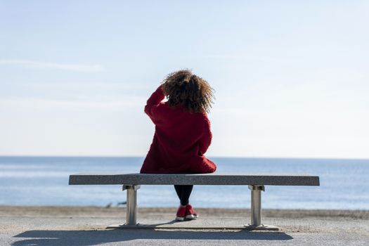 Rear view of a young curly woman wearing red denim jacket sitting on a bench while looking away to horizon over sea