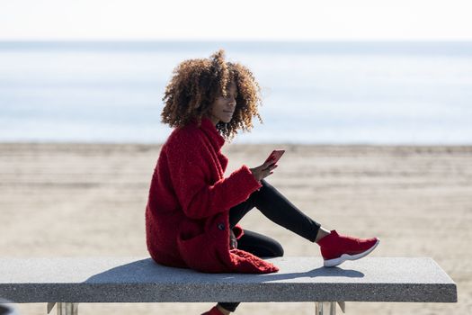 Afro american woman sitting on shore using a mobile phone in a sunny day