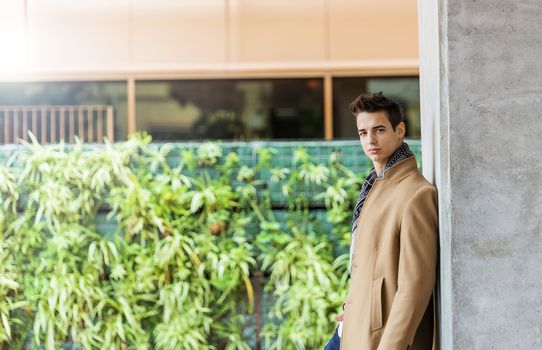 Side view of young trendy man wearing denim clothes leaning on a wall while looking camera outdoors in sunny day