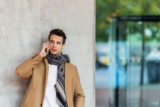 Front view of fashionable young man wearing denim clothes leaning on a wall while using a mobile phone outdoors