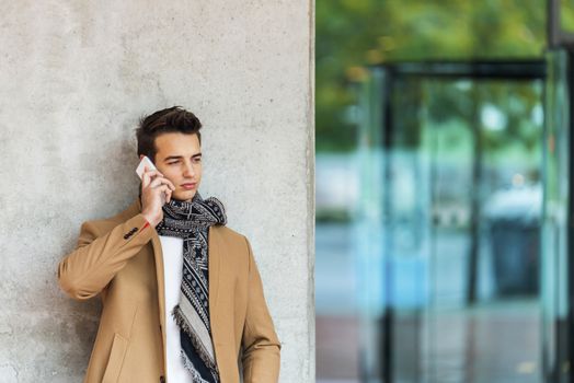 Front view of fashionable young man wearing denim clothes leaning on a wall while using a mobile phone outdoors