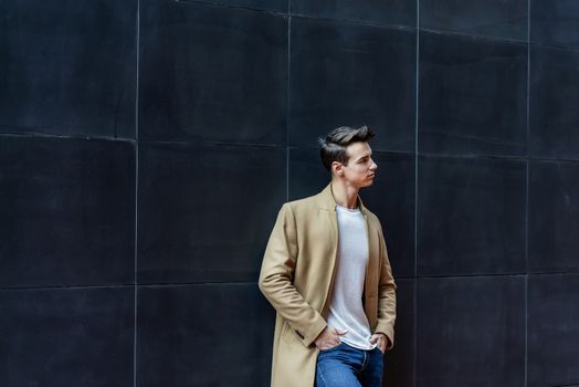 Front view of a trendy young man standing against black wall while looking away outdoors in the street