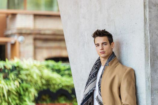 Side view of young trendy man wearing denim clothes leaning on a wall while looking camera outdoors in sunny day