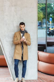 Front view of fashionable young man wearing denim clothes leaning on a wall while using a mobile phone outdoors