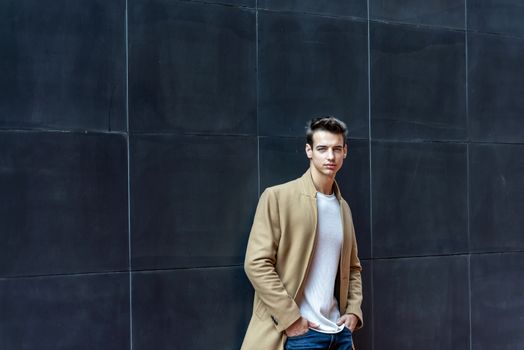 Front view of a trendy young man standing against black wall while looking camera outdoors in the street