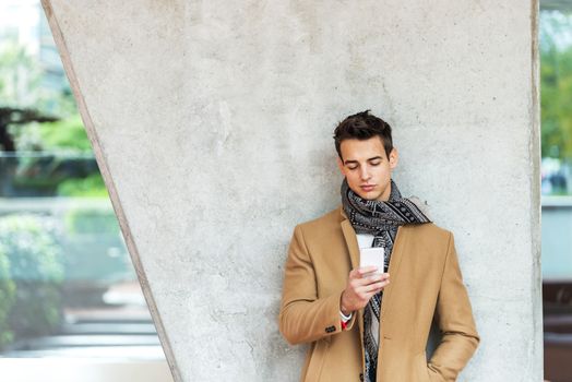 Front view of fashionable young man wearing denim clothes leaning on a wall while using a mobile phone outdoors